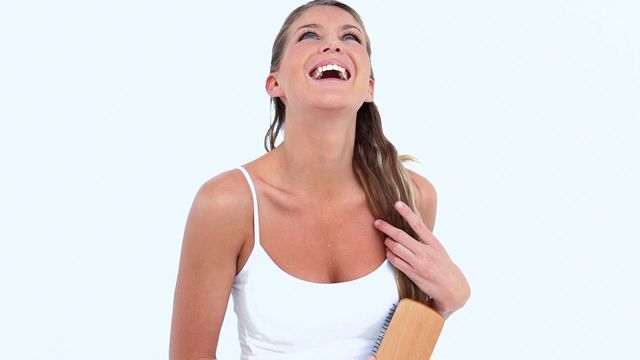 Captures a woman wearing a white tank top, joyfully brushing her long hair against a plain white background. The image projects a sense of carefree happiness. Ideal for use in beauty or wellness content, articles on self-care routines, or product promotions for hair care products.