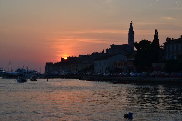 Image shows a picturesque sunset over a historic coastal town, featuring charming waterfront buildings and a distant church steeple. The vibrant sky and the tranquil waters create a serene atmosphere, while the silhouettes of boats add to the charm. Ideal for use in travel brochures, blog posts about coastal vacations, architecture sites, or sunset-themed backgrounds.