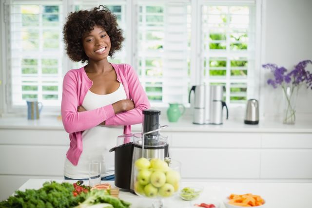 A woman stands in a modern kitchen with arms crossed, smiling confidently. She is surrounded by fresh vegetables and fruits, with a juicer prominently placed on the counter. This image is ideal for promoting healthy living, kitchen appliances, cooking blogs, and home lifestyle content.