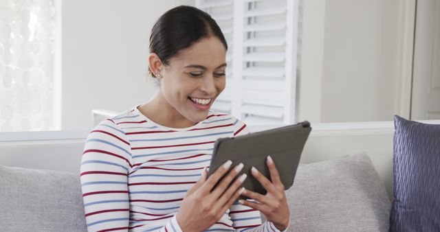 Young woman enjoying using digital tablet while sitting on a couch at home. Perfect for topics related to technology, lifestyle, leisure, home environment, and casual comfort. Ideal for promotional materials, editorial content, and advertising in digital and print media.