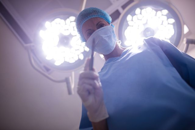 Surgeon in blue scrubs and mask performing an operation under bright surgical lights in a hospital operating room. Ideal for use in medical articles, healthcare websites, educational materials, and promotional content for hospitals and medical services.
