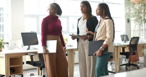 Diverse female coworkers collaborating in modern open office holding laptops and tablet