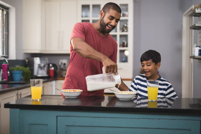 Father pouring milk into son's cereal bowl in a modern kitchen. Both are smiling and enjoying the moment. Ideal for use in family-oriented advertisements, parenting blogs, and healthy lifestyle promotions.