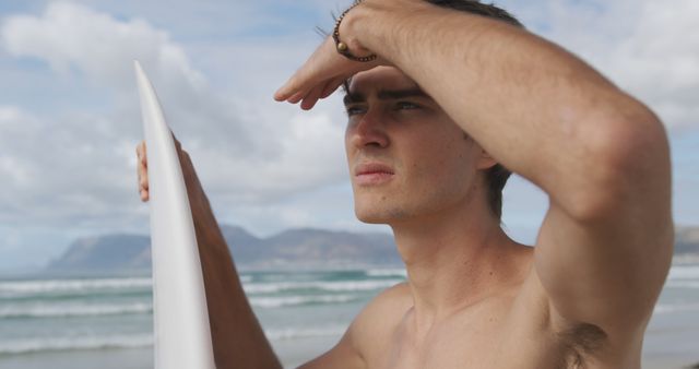 Young man holding surfboard while gazing at ocean horizon. Ideal for use in articles and advertisements about surfing, summer vacations, beachfront activities, and adventure sports. Visually represents anticipation, nature, and active, beach lifestyles.