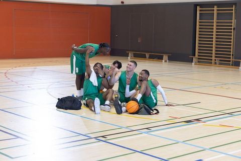 Basketball team sitting and laughing on indoor gym court with ball and backpacks
