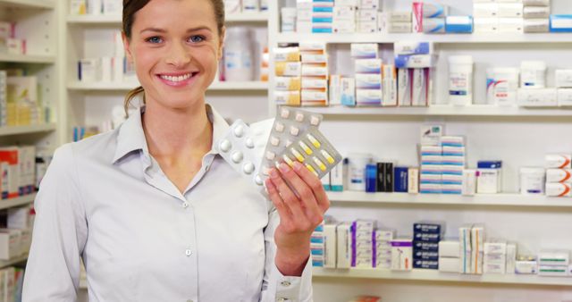 Pharmacist standing in pharmacy, holding blister packs of pills with a smile, surrounded by shelves stocked with various medicines. This image can be used for healthcare-related advertisements, articles on pharmacy services, or promotions of friendly customer service in drugstores.