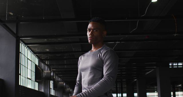 Young man with athletic build wearing grey shirt, standing in modern gym with focused expression. Ideal for fitness, training, health promotion, gym advertisements, and workout inspiration materials.