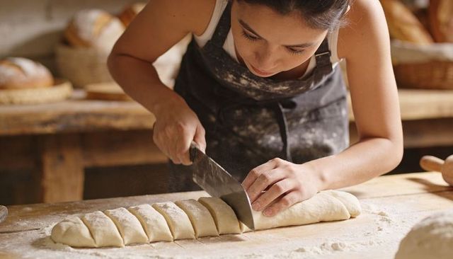 Female baker slicing artisan dough on rustic wooden workbench, flour-dusted apron