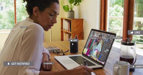 Woman taking notes during online tutorial at home desk with laptop coffee and greenery