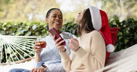 Friends laughing on hammock with smartphone and credit card, wearing santa hat