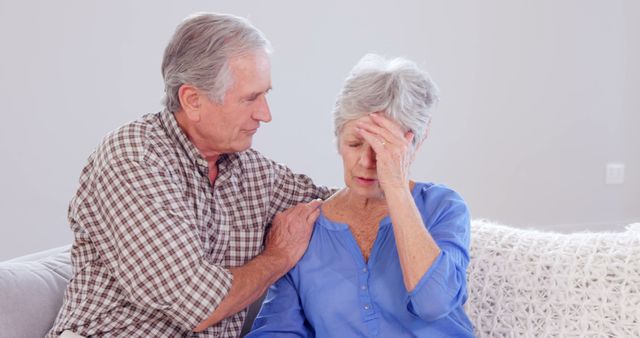 Elderly man placing hand on shoulder of distressed woman, both seated at home. Scene depicting support and concern between senior couple. Ideal for concepts of elder care, relationships, emotional support, and family bonds.