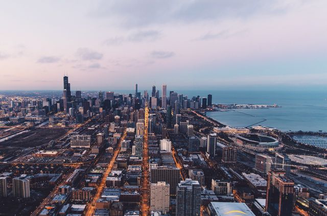 Aerial shot capturing Chicago's skyline at dusk, showcasing illuminated city lights, tall skyscrapers, and the tranquil lakefront. Ideal for urban tourism advertising, real estate promotion, business presentations, and travel blogs showcasing Chicago's iconic landmarks and vibrant city life.