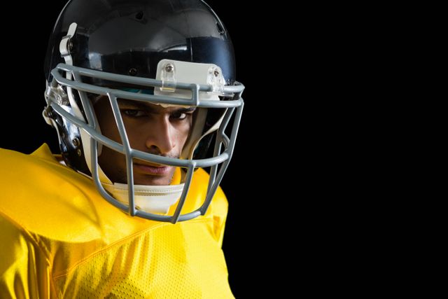 Close-up of an American football player wearing a yellow jersey and helmet, looking intensely at the camera. Ideal for use in sports promotions, team posters, athletic advertisements, and articles about football. The black background emphasizes the player's focus and determination.