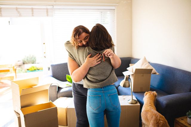 Lesbian couple embracing in living room surrounded by moving boxes, symbolizing new beginnings and domestic lifestyle. Ideal for use in articles or advertisements about relationships, moving, home life, LGBTQ community, and family bonding.