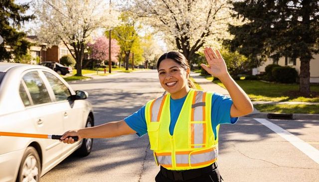 Smiling crossing guard waving traffic baton at sunlit suburban crosswalk in hi-vis vest