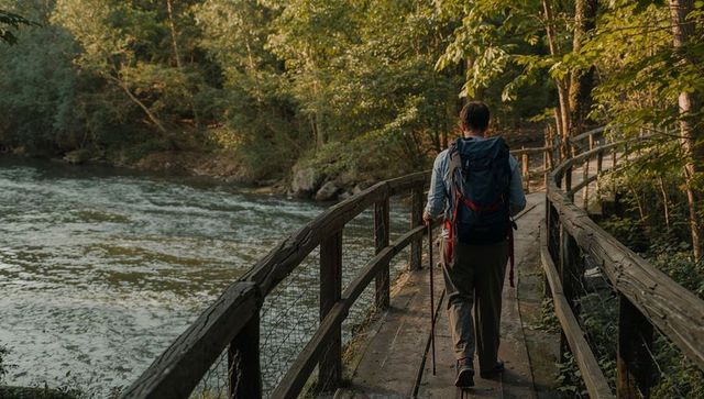 Backpacker walking riverside boardwalk at golden hour with trekking poles and backpack