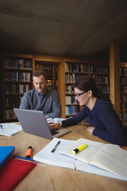 Mature students collaborating on a project in a college library, using a laptop and surrounded by books and notes. Ideal for educational content, adult learning programs, university promotional materials, and articles on lifelong learning and teamwork.