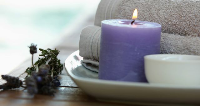 Features lit purple candle and neatly rolled spa towels arranged on a tray. Ideal for promoting spa services, relaxation products, wellness blogs, and self-care routines. Perfect for creating atmosphere of tranquility and comfort.