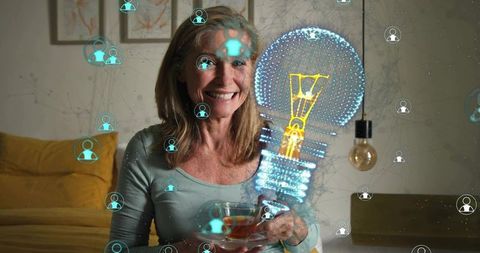 Smiling senior woman holding teacup viewing holographic lightbulb network connecting people