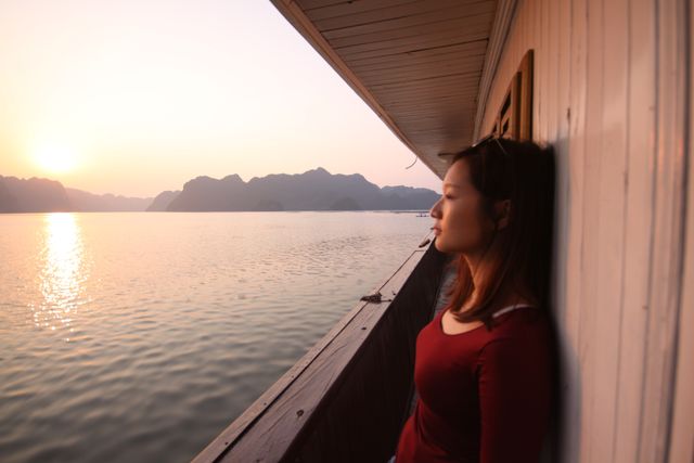 Woman leaning against the side of a boat, enjoying the peaceful scenery during sunset. Background filled with calm waters and distant mountains. Ideal for use in travel promotions, vacation brochures, and advertisements focusing on serene escapes and relaxation.
