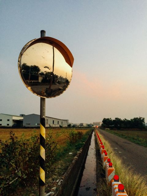 This depicts a convex mirror reflecting a rural road, combined with safety barriers, at dusk. The sky shows the colors of the setting sun, creating a serene countryside landscape. Useful for themes related to road safety, rural infrastructure, and countryside tranquility.