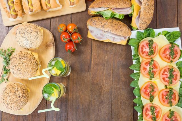 Assorted sandwiches, including burgers and hot dogs, are arranged on a wooden table alongside a fresh salad with tomatoes, basil, and cheese. Two glasses of mojito with straws add a refreshing touch. Ideal for use in food blogs, summer party invitations, picnic promotions, or healthy meal planning content.