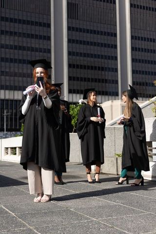 Female graduates taking selfie on urban plaza holding diplomas in caps and gowns
