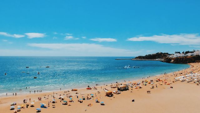 Beach scene features numerous people sunbathing, swimming, and enjoying leisure activities under a bright summer sky. Sandy shore dotted with umbrellas and beachgoers. Calm ocean extends to the horizon with coastal landscapes in the background. Ideal for tourism advertisements, travel brochures, summer vacation promotions, and coastal destination features.