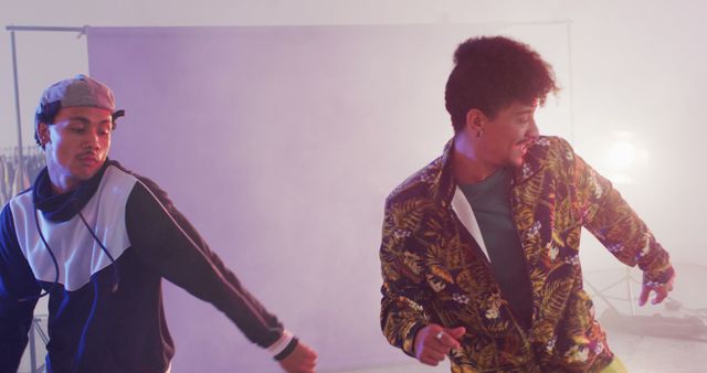Two young men are dancing energetically in a studio, filled with joy and enthusiasm. One is wearing a patterned jacket, the other a sporty outfit. Smoke and soft colored lights are seen in the background, creating a vibrant atmosphere. Suitable for use in content related to dance, youth culture, urban lifestyles, and entertainment.