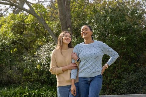 Diverse women walking arm-in-arm in sunlit garden smiling, enjoying outdoor stroll