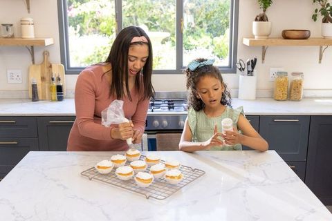 Diverse mother and daughter decorating cupcakes together on marble kitchen island at home