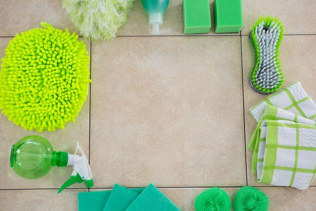 Green cleaning products arranged on a tiled floor, showcasing various eco-friendly cleaning supplies such as sponges, a scrub brush, a spray bottle, and microfiber cloths. Ideal for use in articles or advertisements promoting eco-friendly cleaning solutions, household hygiene, and sustainable living.