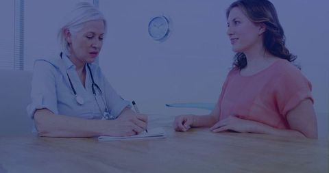 Female doctor writing patient notes during consultation with woman in coral top