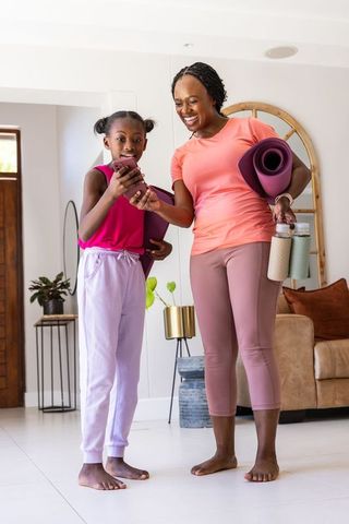 African mother and daughter preparing for home yoga, checking smartphone while carrying mats