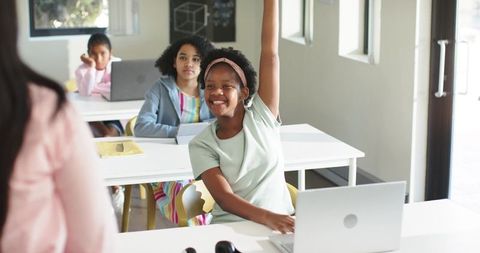 Smiling African American schoolgirl raising hand in modern classroom with laptop