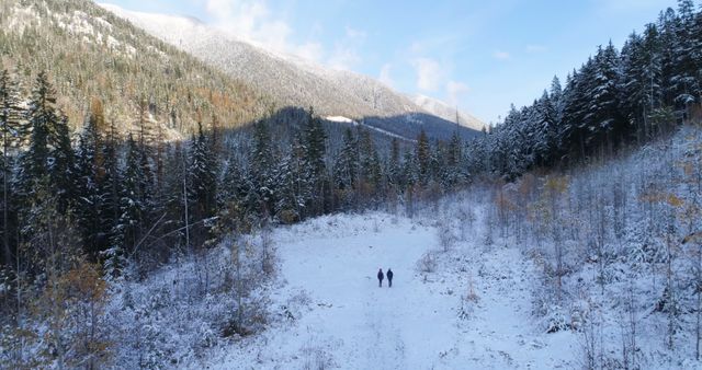 Two people are seen walking through a snow-covered clearing surrounded by forested mountains, with copy space. Their journey amidst the serene winter landscape emphasizes the beauty and tranquility of nature.