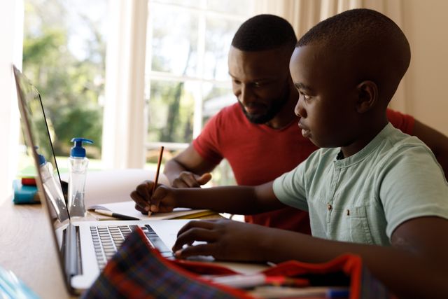 Side view of an african-american boy studying on a laptop with his father beside him helping. on the table beside the laptop is a hand sanitizer.