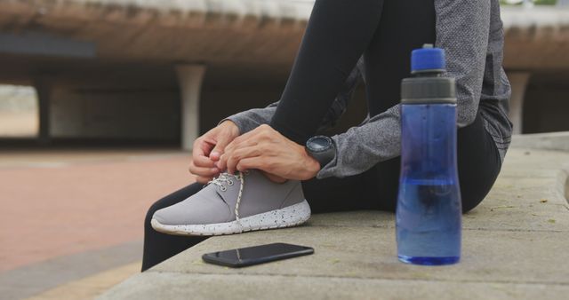 The image shows a person tying their shoe laces on a concrete surface before exercising. Nearby are a water bottle and smartphone, suggesting preparation for a workout. This image can be used for fitness blogs, workout plans, healthy lifestyle promotions, and sportswear advertisements.