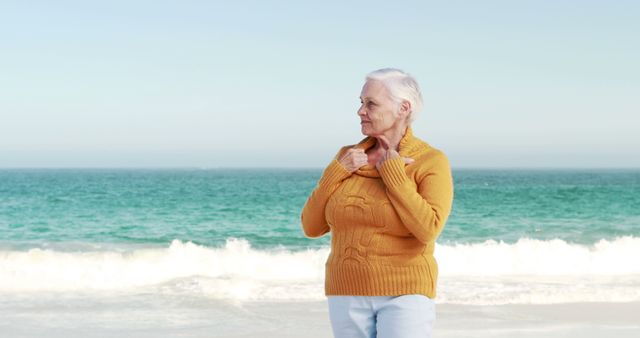 Senior woman in yellow sweater standing on sandy beach with ocean waves in background, enjoying peaceful moment. Useful for ads promoting senior lifestyle, relaxation, retirement, travel, health products targeting the elderly.