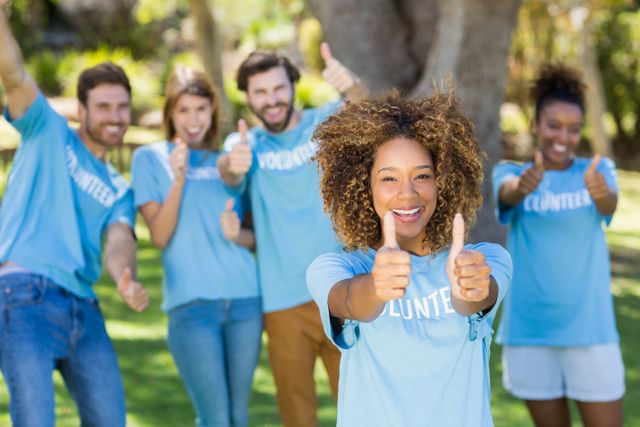 Diverse group of volunteers wearing blue shirts giving thumbs up in a park. Ideal for promoting community service, teamwork, charity events, social work, and unity. Perfect for use in advertisements, brochures, websites, and social media campaigns focused on volunteerism and community engagement.