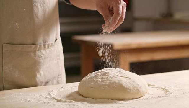 Baker Sprinkling Flour Over Risen Dough on Rustic Wooden Countertop