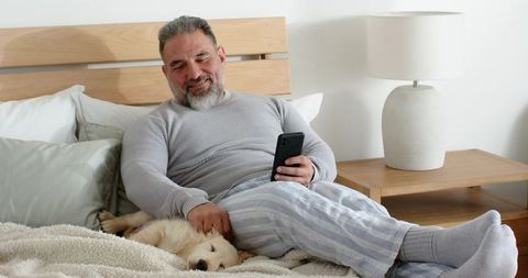 Mature man relaxing in bed petting puppy while using smartphone in cozy bedroom