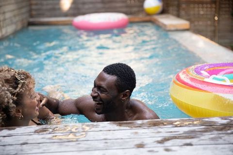 Couple laughing and splashing in backyard pool with rainbow-striped donut float