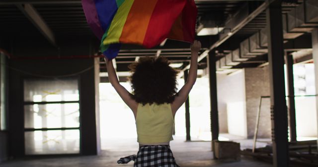 Young woman with curly hair waving rainbow flag in an urban environment, symbolizing pride and celebration of LGBTQ community. Great for promoting diversity, inclusion, and equality. Suitable for social media campaigns, pride events, and awareness content.