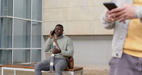 African American man talking on phone and holding takeaway cup on urban bench with scarf