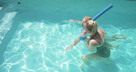 Female instructor teaching child swimming with foam noodle in sunlit turquoise pool