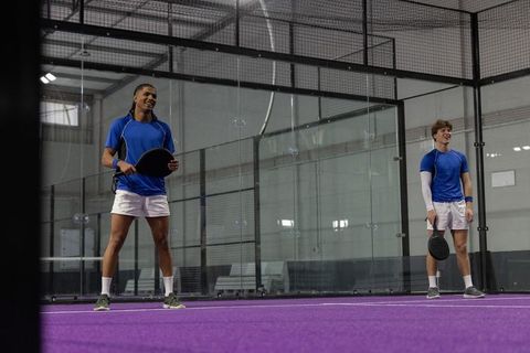 Diverse male padel doubles smiling and standing on vibrant purple indoor court, team unity