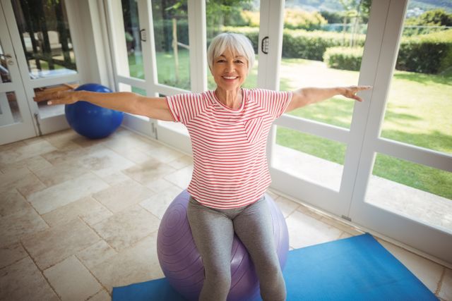 Senior woman stretching on a fitness ball in a bright, spacious room with large windows. Ideal for promoting healthy aging, home exercise routines, and active lifestyles. Perfect for use in articles, blogs, and advertisements focused on senior fitness, wellness, and home workout programs.