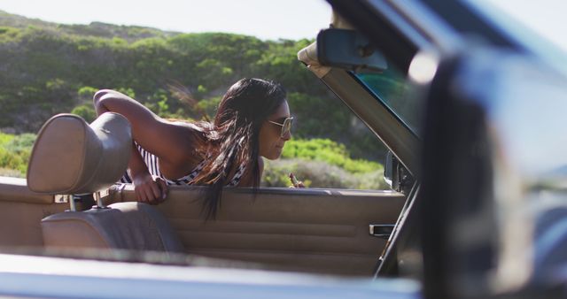 African american woman applying lipstick while looking in the mirror of convertible car on road. road trip travel and adventure concept