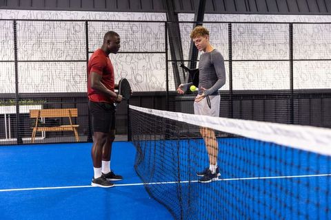 Two male padel players training and coaching at net on vibrant blue indoor court